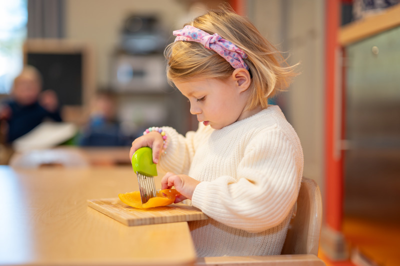 A toddler student sitting at a table cutting a pepper.